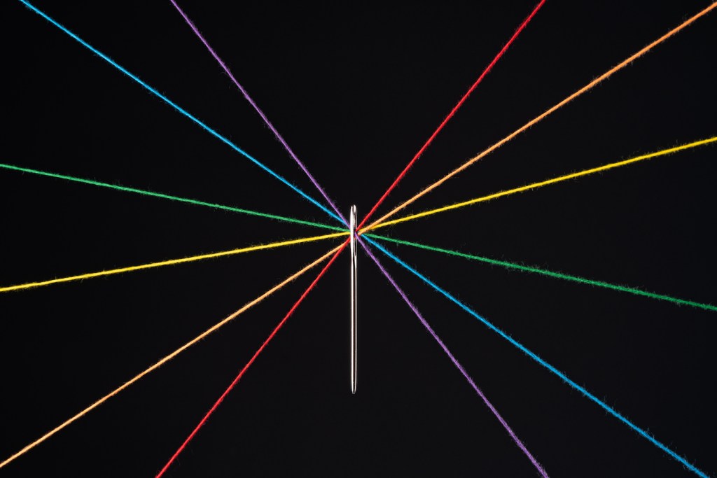 Rainbow Colored Threads Through Needle Eyelet Against Black Colored Background.