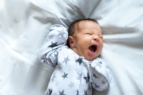 Top view of newborn baby lying on bed and crying or shouting or yawning. Infant lying on white bed.