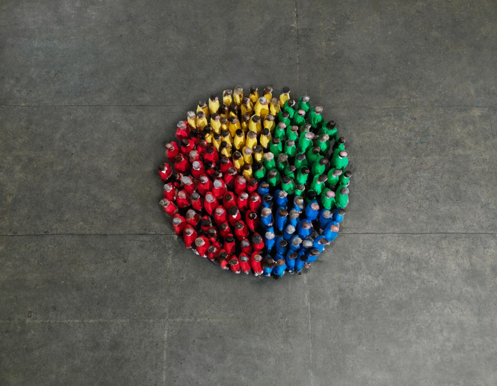 A crowd of people wearing red, yellow, green and blue coloured shirts, forming a pie chart shape; fractional hiring for startups, hiring contractors