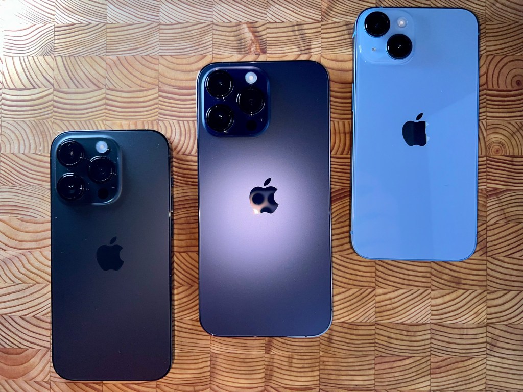 iPhone 14 pro space black, iPhone 14 pro deep purple and iPhone 14 blue colored iPhones on a wooden surface