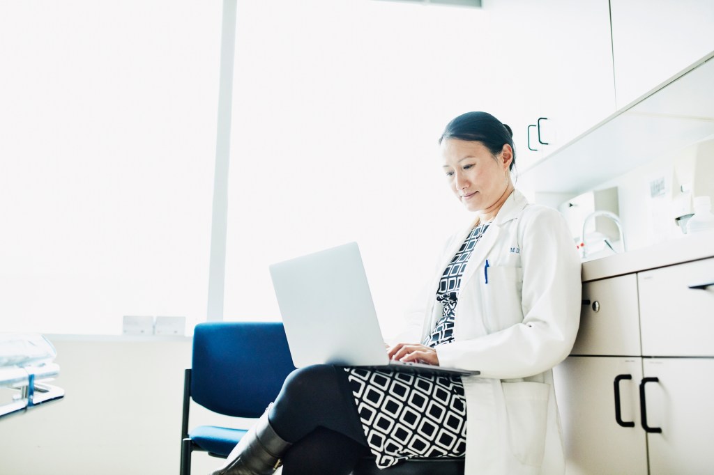 Portrait of female doctor working on laptop in exam room, used in a post about healthcare community Docquity