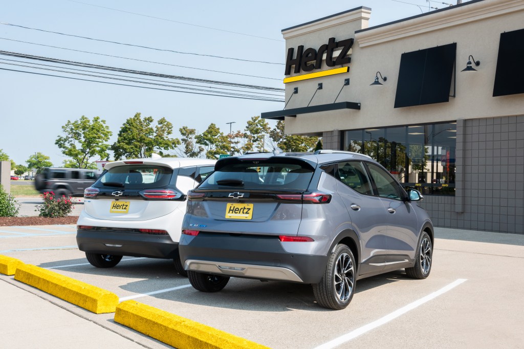 A Summit White 2023 Chevrolet Bolt EV and a Gray Ghost Metallic 2023 Chevrolet Bolt EUV parked in front of a Hertz rental location in Michigan. Hertz will begin taking deliveries of the Chevrolet Bolt EV and Bolt EUV in the first quarter of 2023.