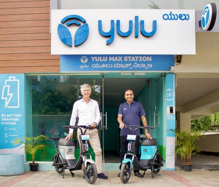 Two men with e-bikes and helmets in front of shop front that says Yulu