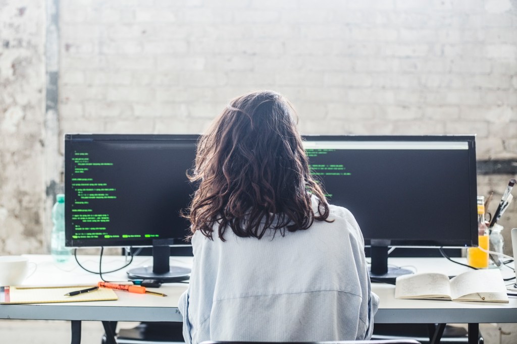 back of woman sitting at desk with two monitors