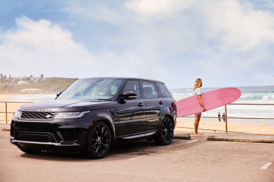 a surfer holds a pink surfboard standing behind a black SUV near the beach