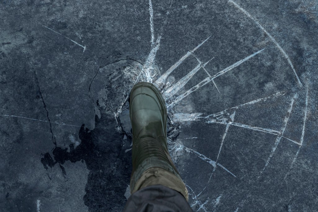 Fisherman foot on broken cracked thin ice at lake. Dangerous fishing