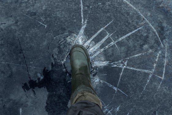 Fisherman foot on broken cracked thin ice at lake. Dangerous fishing