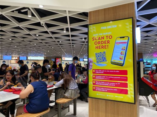 Food court scene, with people seated at tables eating and a sign with details about ordering via a QR code.