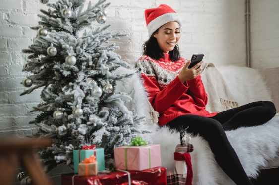 girl, wearing a Santa hat and holding a cell phone, sitting on a couch next to a Christmas tree