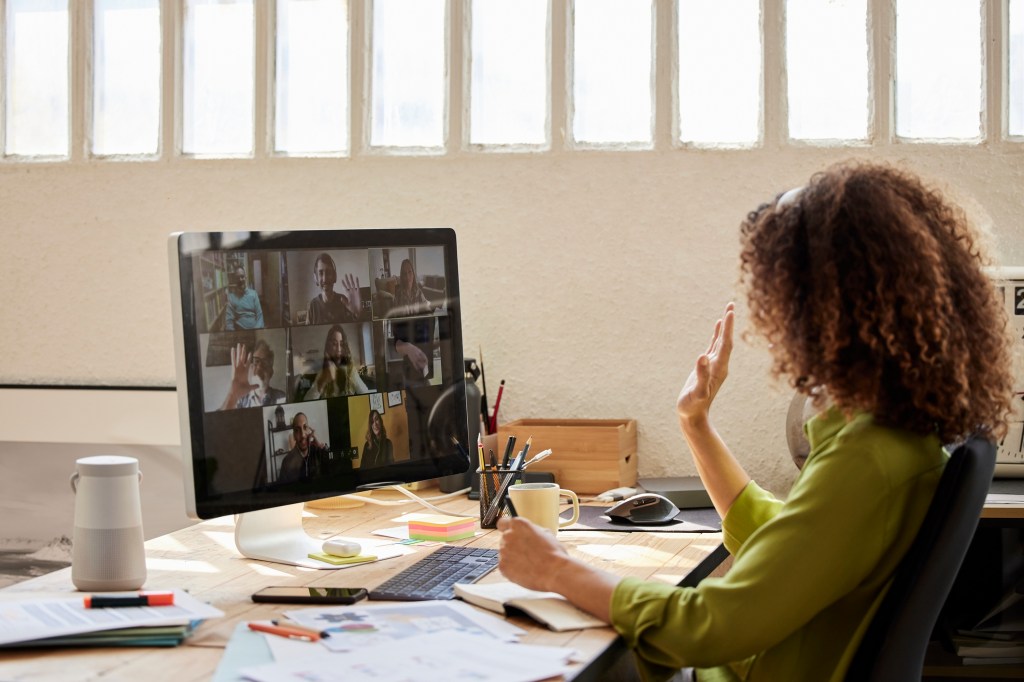 Female freelancer waving hand with colleagues while attending video call at home office. Businesswoman with coworkers planning strategy. They are discussing together.
