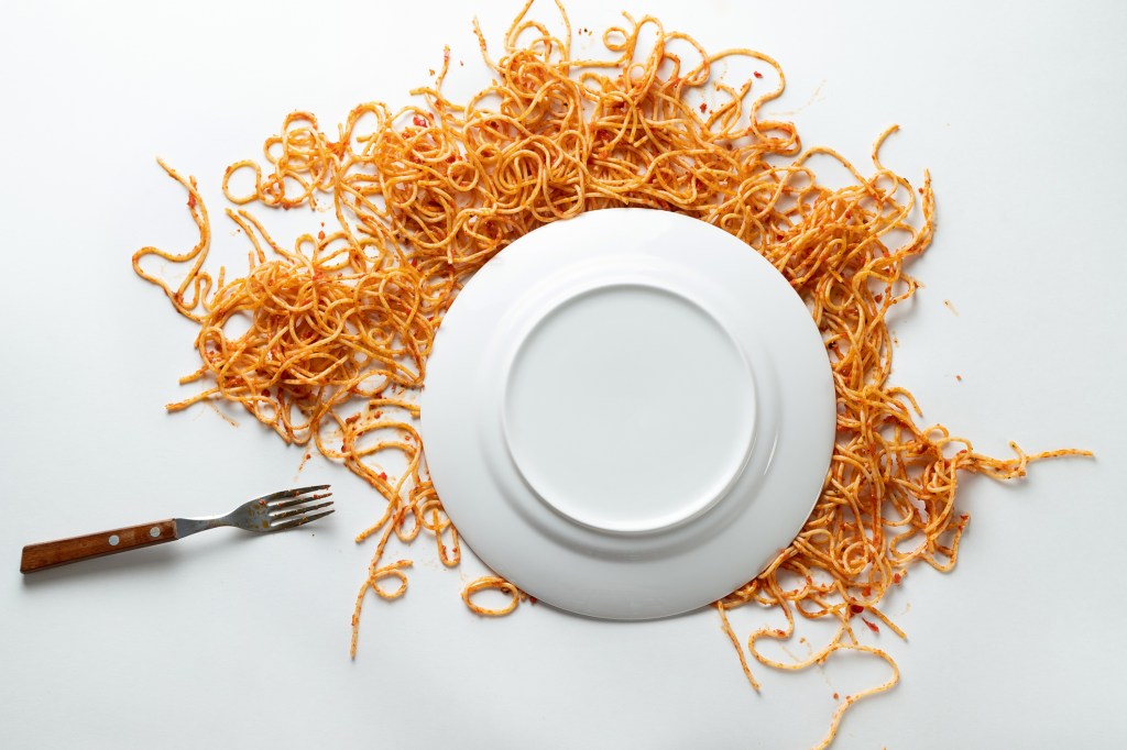 A fallen white ceramic plate of spaghetti, with a fork beside it. Pasta bolognese in tomato sauce is scattered on a white background or table. The concept of vegetarian and vegan food. Food background. Copy of the text space.