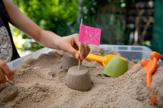 Child placing a homemade flag made from a pink sticky note with a drawing of a smiley face, into the top of a small sandcastle surrounded by sand inside a plastic sandbox.