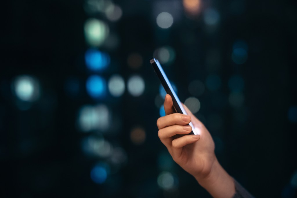 Close up shot of female hand using smartphone at night, against illuminated and defocused bokeh in background.