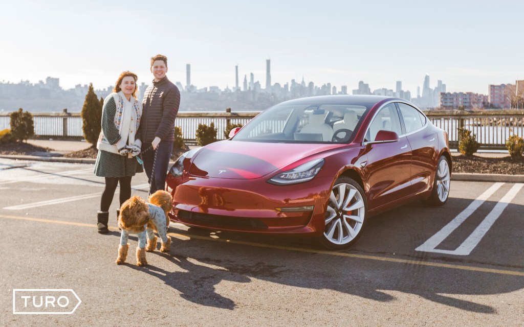 man woman and dog standing in front of red tesla in a parking lot