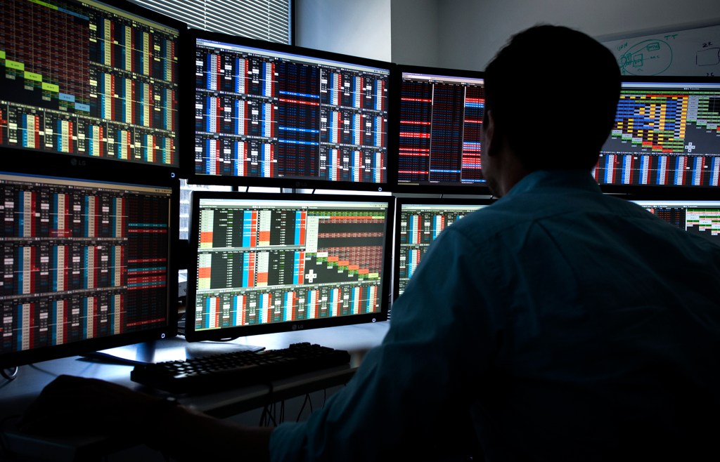 shadowy figure sitting in front of a set of computer monitors using X_Trader software in a darkened room