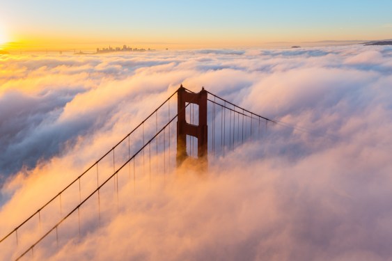 One of the towers of the Golden Gate Bridge juts up through fog at sunrise.