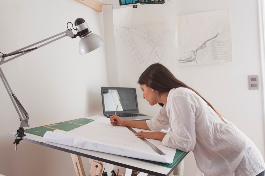 Female architect working at a drafting table with a laptop open beside her
