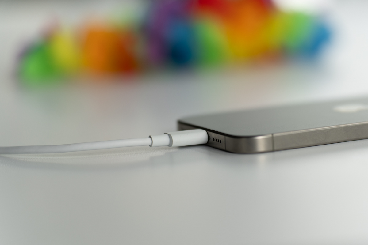 The natural titanium iPhone 15 Pro Max, face down on a table, showing the bottom of the phone and an attached USB-C cable