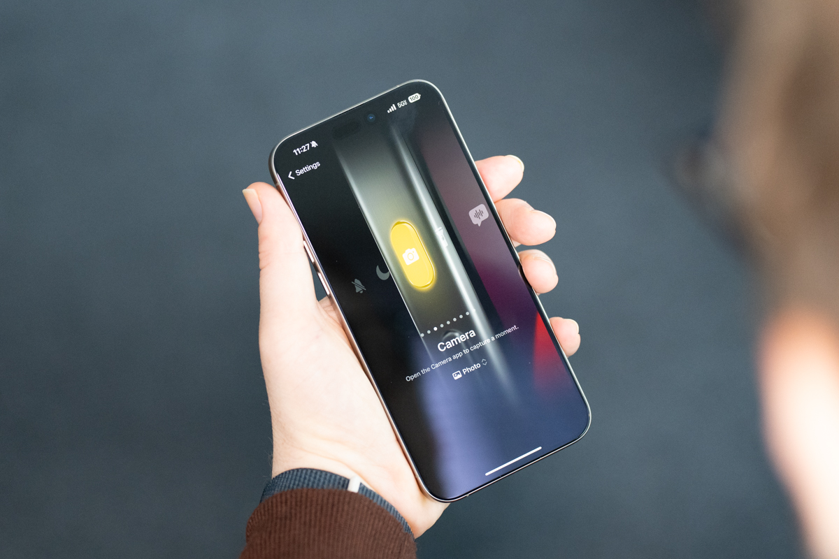 iPhone 15 Pro Max in natural titanium, being held, showing the display and Action button customization interface