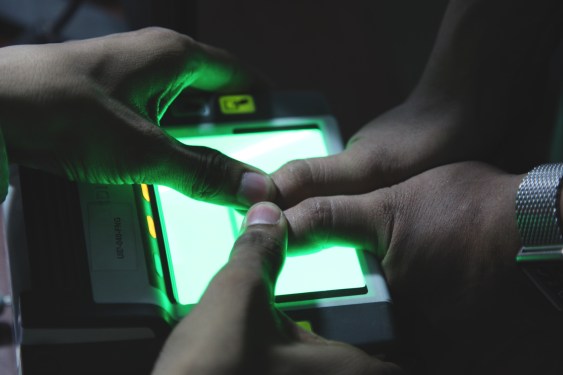 a person holding down the thumbprints of an Indian citizen having their biometrics taken on a device with a bright glowing green light