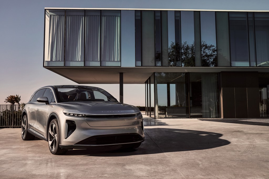 A promotional shot shows a silver Lucid Gravity SUV parked in front of a modern house with big glass windows.