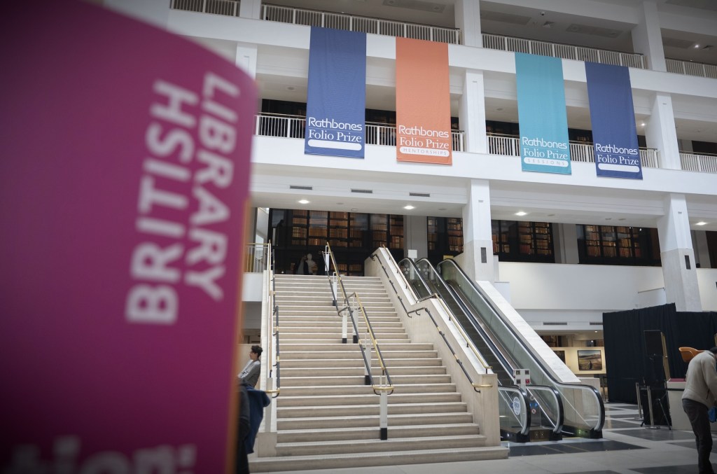 an outside photo of the British Library featuring a sign in purple in the foreground and in the background the outer exterior of the British Library building in London