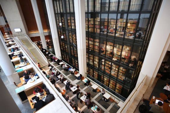 an angled shot looking down at the inside of the British Library, with desks on the ground floor and a huge glass wall with books behind
