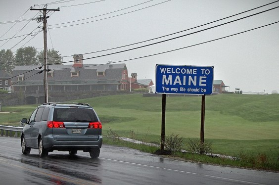 a blue roadsign that says "Welcome to Maine, the way life should be" on a road with a car driving past, on a dark and slightly rainy day with a large field and house in the background.