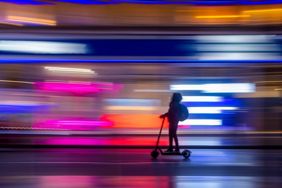A stand up electric scooter passes brightly lit colourful shop fronts at night on New Oxford Street in London's West End leaving light trails.
