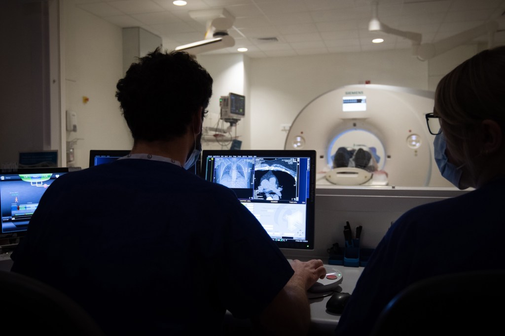 a photo of a person having a CT-scan in the background, with two radiographers behind protective glass in front of the CT scanner's computer