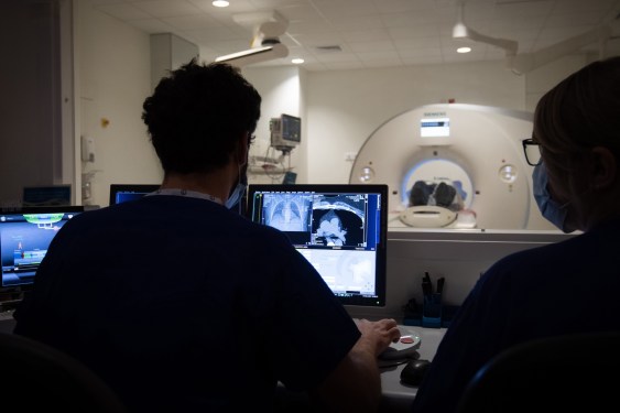 a photo of a person having a CT-scan in the background, with two radiographers behind protective glass in front of the CT scanner's computer