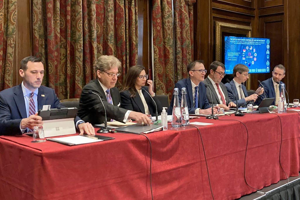 a photo of UK and US officials sitting at a red table discussing the LockBit takedown