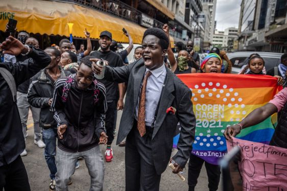 Man walking and shouting in front of rainbow colored protest sign with his arm in the air