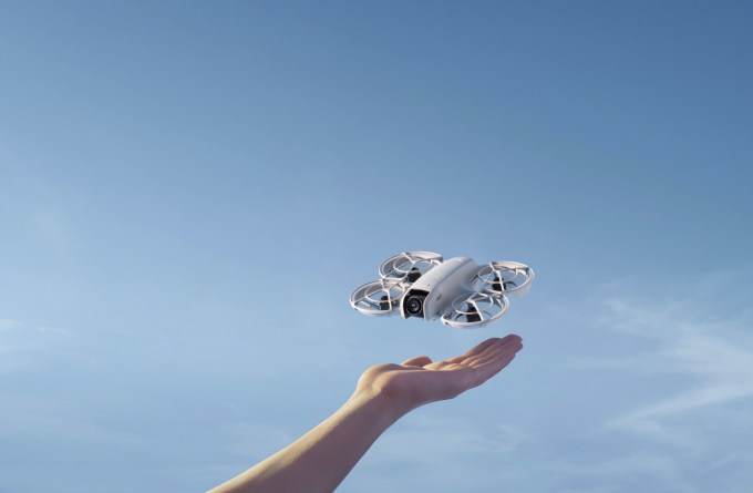 a forearm and hand raised up in front of a blue sky. a small white drone is hovering a few inches above the hand