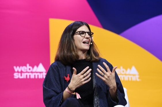 A brown haired woman wearing glasses stands in front of the a multi-colored background with the web summit logo