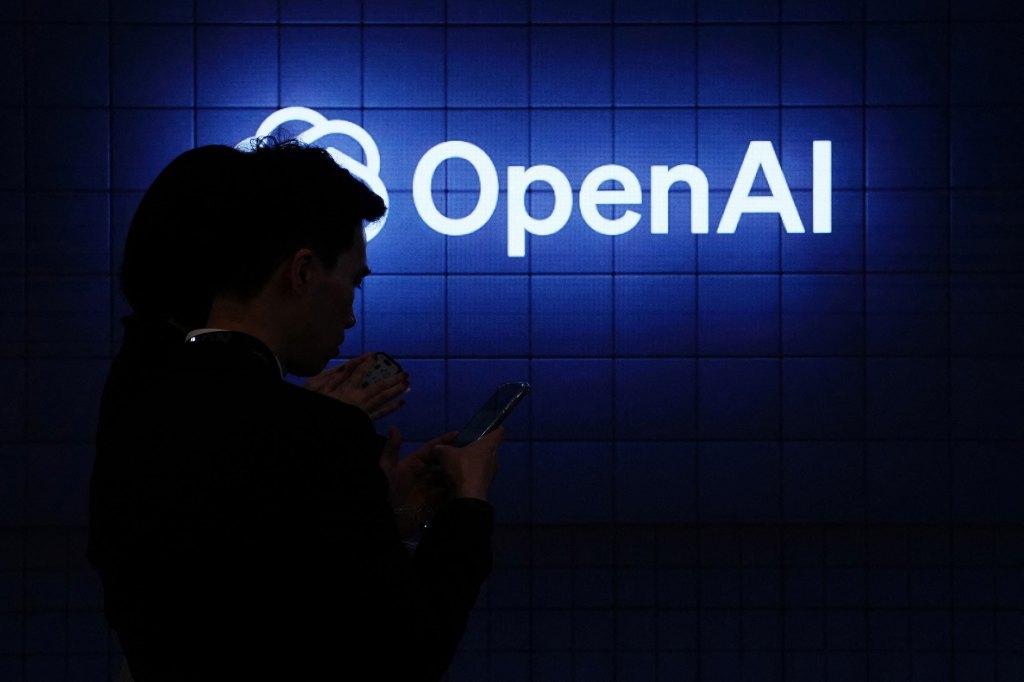 Visitor look at their phone next to an Open AI's logo during the Mobile World Congress (MWC), the telecom industry's biggest annual gathering, in Barcelona on February 26, 2024. The world's biggest mobile phone fair throws open its doors in Barcelona with the sector looking to artificial intelligence to try and reverse declining sales. (Photo by PAU BARRENA/AFP via Getty Images)